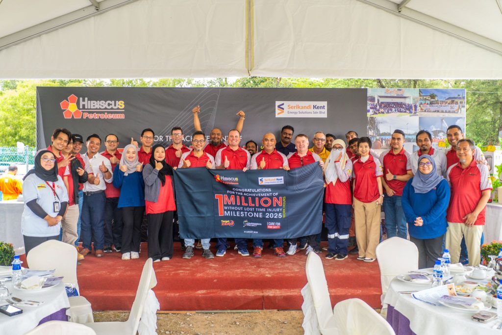 A large group of people wearing red and white shirts pose together under a tent, smiling and giving thumbs up while holding a banner. The banner reads: “Congratulations for the achievement of 1 million without lost time injury milestone 2025” with logos of Hibiscus Petroleum and Serikandi Kent Energy Solutions Sdn Bhd. The background includes the companies’ logos on a display board, and tables with white tablecloths are set in the foreground.